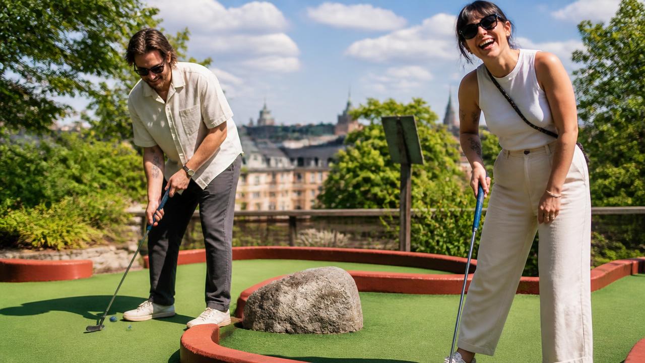 Två personer spelar minigolf i solsken med Stockholms hustak i bakgrunden